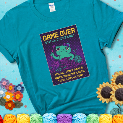 Teal t-shirt shows a pixel-style design of a grumpy yarn frog holding a hook, surrounded by yarn chaos, with the text “Game Over. Stitch Count Lost.”