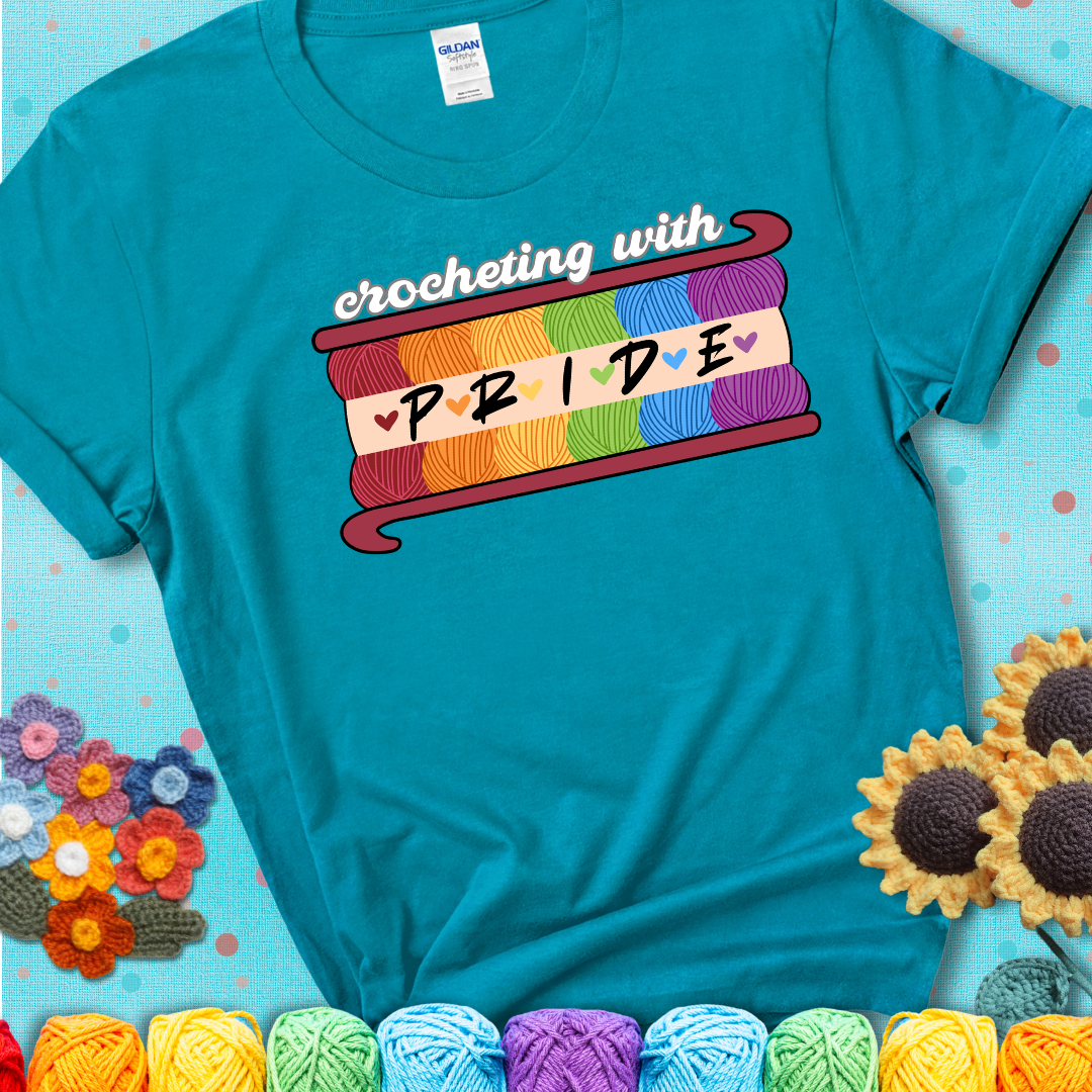 Yarn skeins in rainbow colors stacked in a hook shape. Text reads “Crocheting With Pride” showing a blue green teal tee with an aqua sunflower background