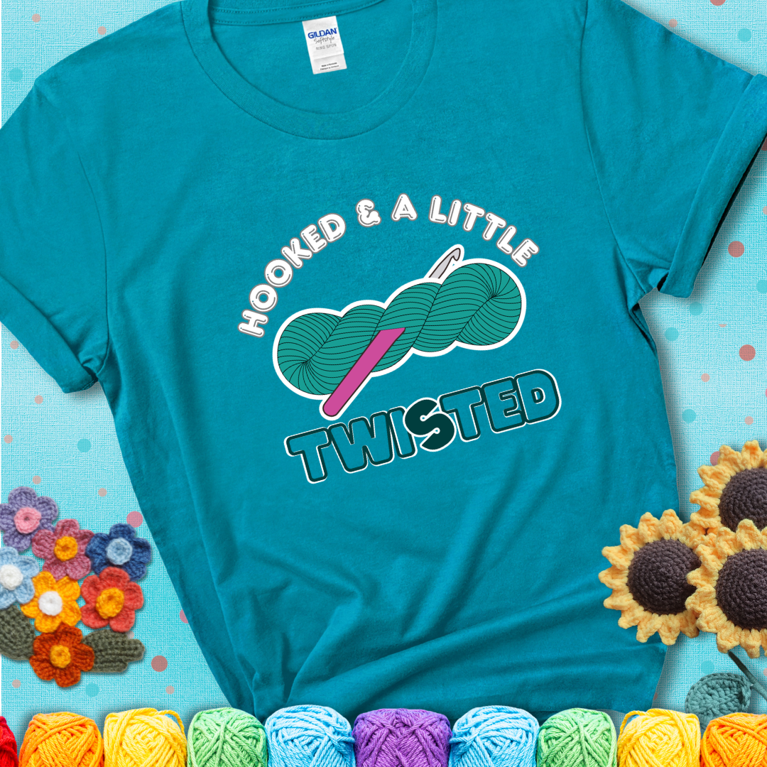 A teal skein of yarn with a hot pink crochet hook through it, captioned “Hooked & a Little Twisted” on a teal t-shirt.