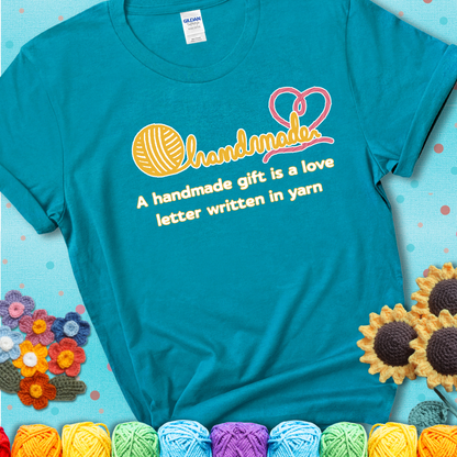 Soft script-style design with heart detail. Text reads: “A Handmade Gift Is a Love Letter Written in Yarn.” A sweet and sentimental shirt for crocheters who craft from the heart, showing a blue-green teal tee with an aqua sunflower background