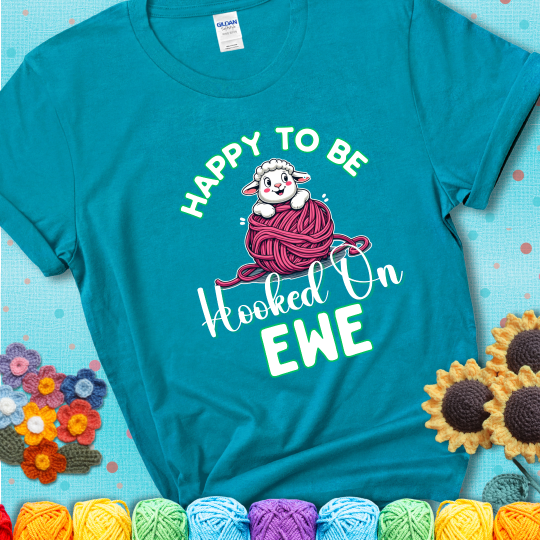 Teal t-shirt with smiling cartoon sheep peeks from a pink yarn ball, surrounded by green text “Happy to Be Hooked on Ewe”, a playful and affectionate sheep pun for crocheters.