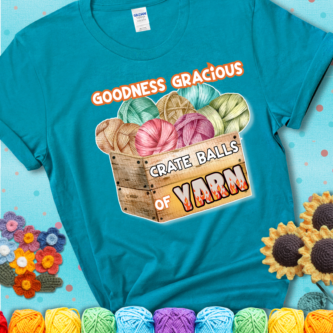 Wooden crate filled with colorful yarn balls and bold text “Goodness Gracious, Crate Balls of Yarn” with flame effects, a funny gift for crocheters showing a blue-green teal tee with an aqua sunflower background