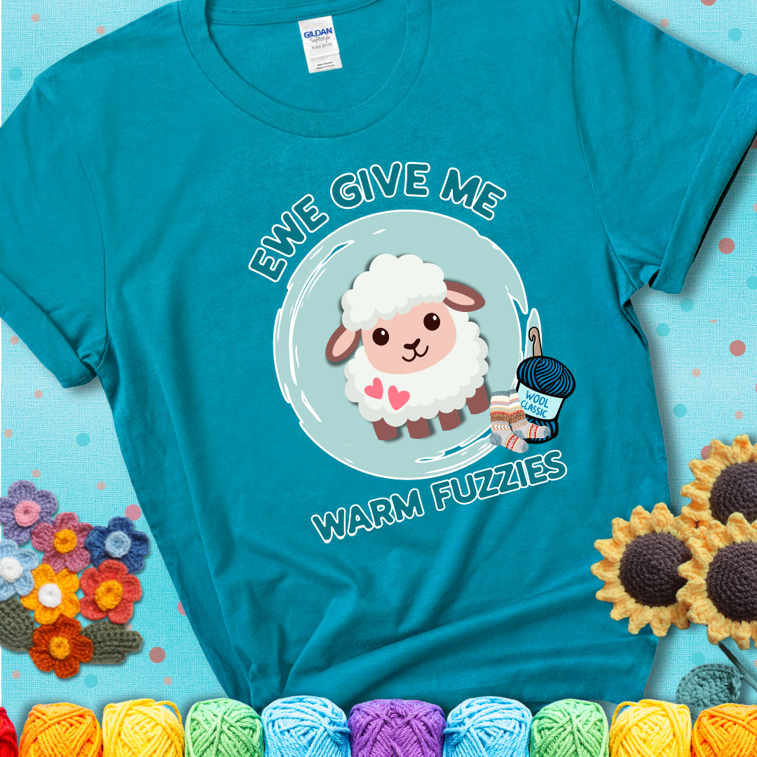 Teal t-shirt showing a cartoon sheep with pink hearts, wool socks, and yarn labeled “Wool Classic.” Text reads “Ewe Give Me Warm Fuzzies.”