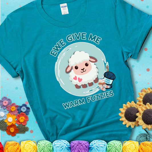 Teal t-shirt showing a cartoon sheep with pink hearts, wool socks, and yarn labeled “Wool Classic.” Text reads “Ewe Give Me Warm Fuzzies.”