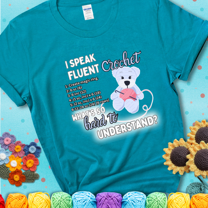 A cute crochet bear holding a yarn ball, surrounded by a short crochet pattern. The phrase reads “I Speak Fluent Crochet – What’s so hard to understand?” on a teal t-shirt.