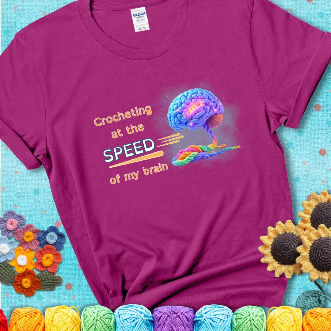 A glowing rainbow brain unravels into colorful yarn with the phrase “Crocheting at the speed of my brain” on a magenta t-shirt. 