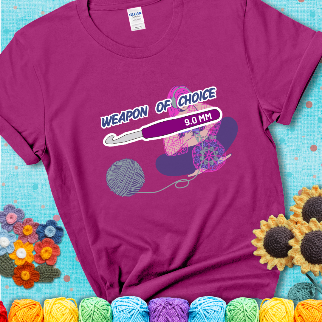 Magenta t-shirt with an Illustration of a crocheter sitting cross-legged with a 9.0 mm crochet hook above her, labeled “Weapon of Choice.” She’s crocheting a colorful mandala-style project from a large yarn ball.