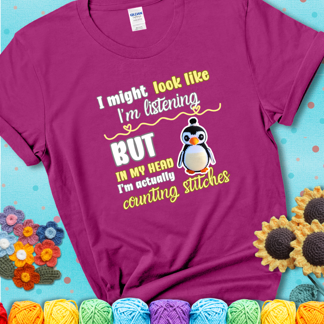 Magenta t-shirt shows a crocheted penguin next to the text “I might look like I’m listening but in my head I’m actually counting stitches.”