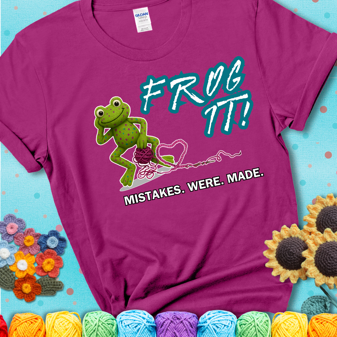 Green frog with yarn and heart-shaped loop. Text reads: “Frog It! Mistakes Were Made.” A playful crochet tee for anyone who's ever had to rip it back, showing a magenta tee with a teal sunflower background
