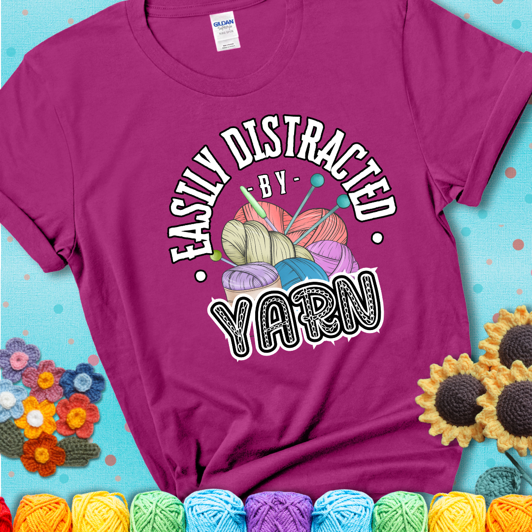 If yarn steals your focus every Design reads 'Easily Distracted By Yarn', perfect crochet gift for yarn lovers; showing a magenta tee with a teal sunflower background