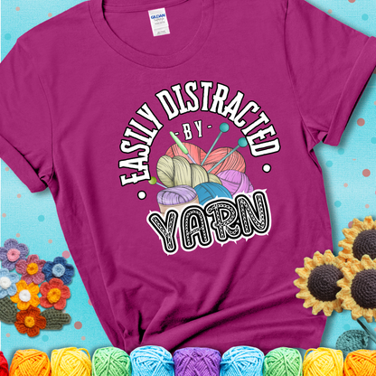 If yarn steals your focus every Design reads 'Easily Distracted By Yarn', perfect crochet gift for yarn lovers; showing a magenta tee with a teal sunflower background