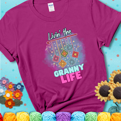 Magenta t-shirt shows colorful crochet granny squares hanging from a hook with text “Livin’ the Granny Life” in bold pink and blue font; a fun nod to classic crochet.