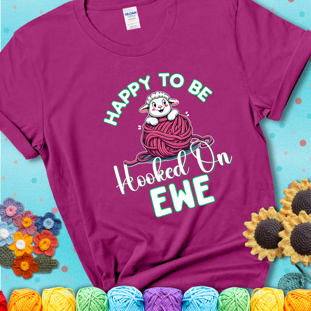 Magenta t-shirt with smiling cartoon sheep peeks from a pink yarn ball, surrounded by green text “Happy to Be Hooked on Ewe”, a playful and affectionate sheep pun for crocheters.