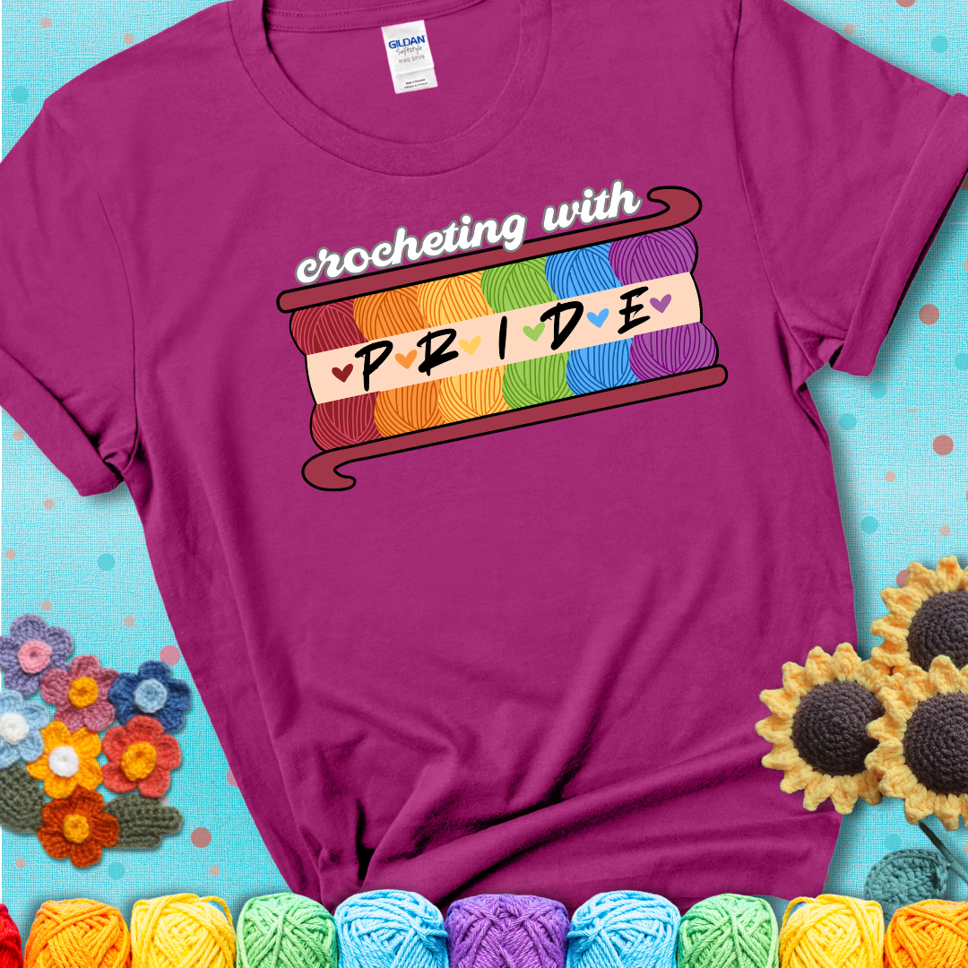 Yarn skeins in rainbow colors stacked in a hook shape. Text reads “Crocheting With Pride” showing a magenta tee with a teal sunflower background