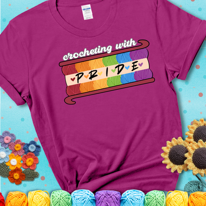Yarn skeins in rainbow colors stacked in a hook shape. Text reads “Crocheting With Pride” showing a magenta tee with a teal sunflower background