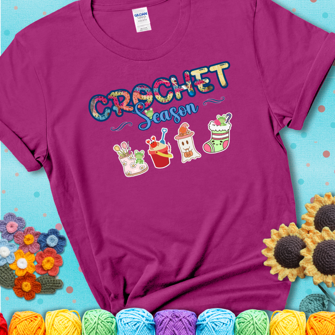Magenta t-shirt shows the words “Crochet Season” with cute seasonal icons below: a spring boot with hooks, summer bucket with crab, fall ghost with pumpkin hat, winter stocking, and tiny frogs.