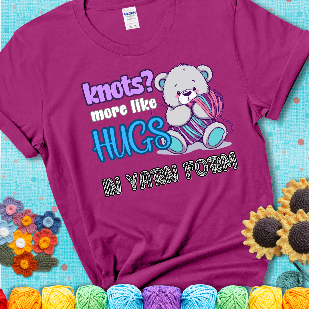 Cute teddy bear holding colorful yarn ball with hearts. Text reads: “Knots? More Like Hugs in Yarn Form.” Perfect for cozy gift-givers and yarn lovers, showing a magenta tee with a teal sunflower background