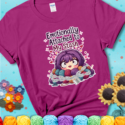 Adorable character with wide eyes hugging a yarn stash wrapped in chains and a lock, with the text “Emotionally Attached to My Stash”  on a magenta tee.