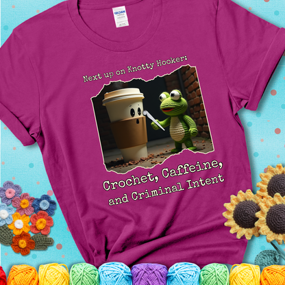 A crocheted frog holding a crochet hook confronts a surprised coffee cup in a dim alleyway with text: "Next up on Knotty Hooker: Crochet, Caffeine, and Criminal Intent," showing a magenta tee with a teal sunflower background