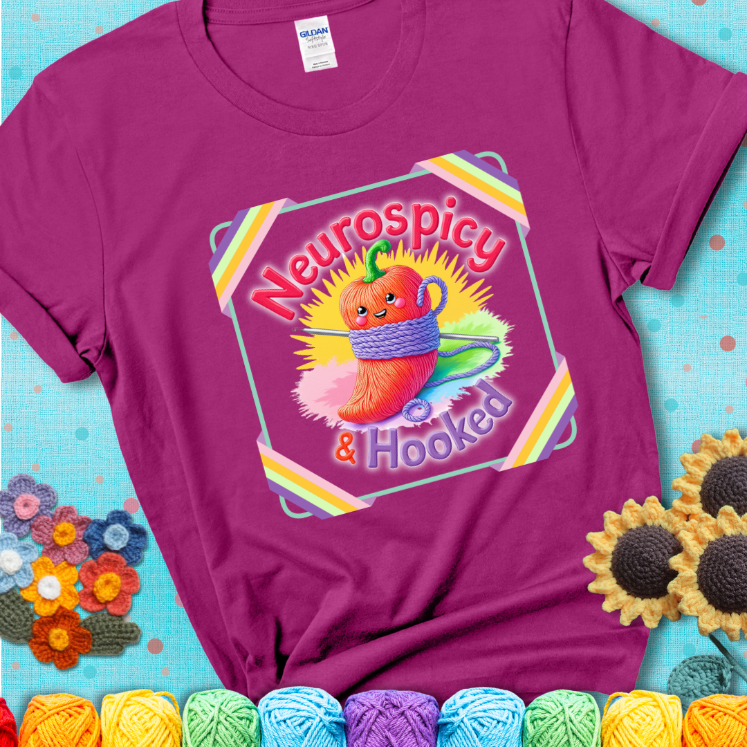 Magenta t-shirt featuring a smiling red chili pepper wrapped in yarn with a crochet hook, text says “Neurospicy & Hooked.”