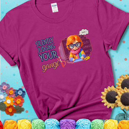 Antique Heliconia t-shirt with a serious-looking crocheter in rainbow clothing and big glasses, holding a tape measure under the phrase “Silently Judging Your Gauge.”