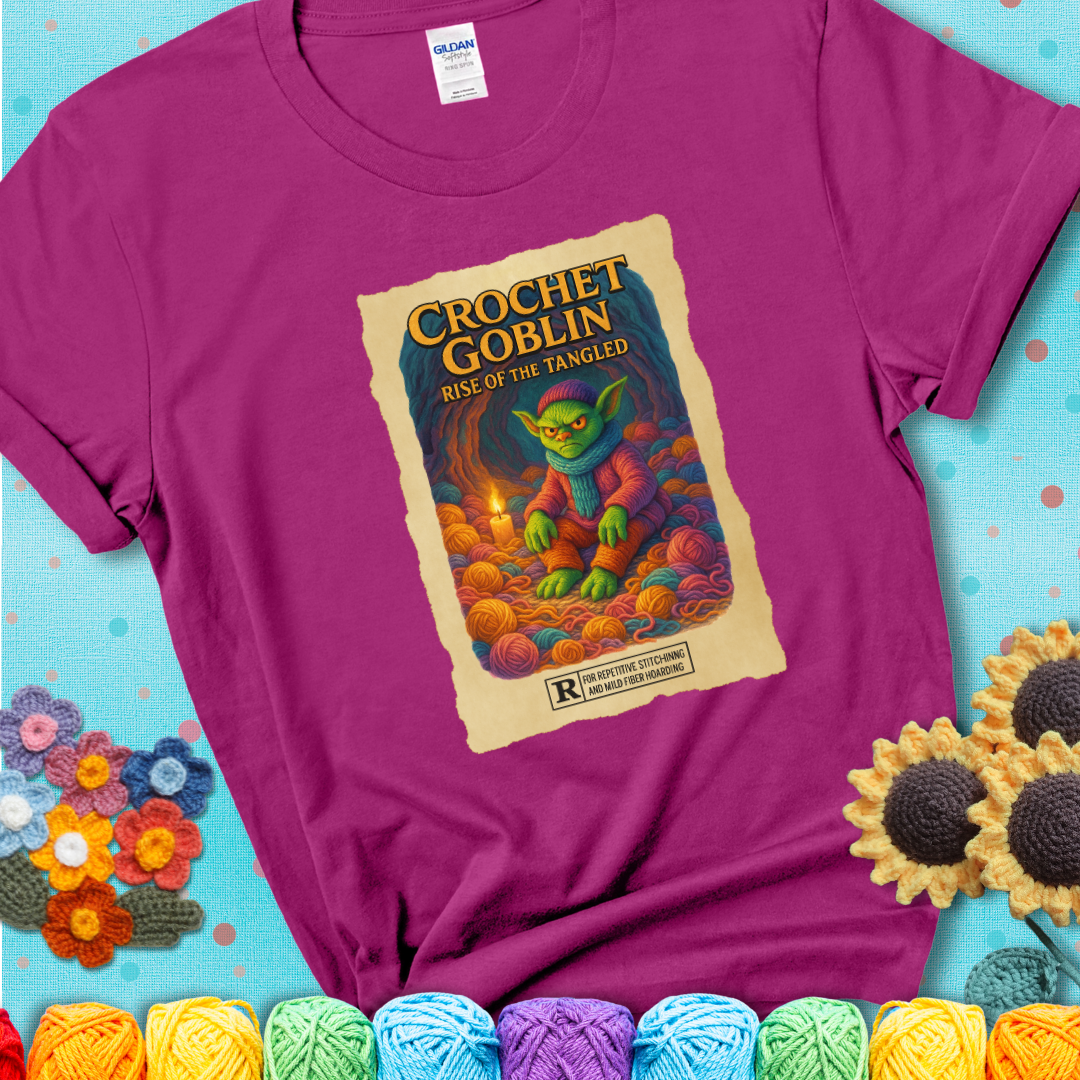 Crochet goblin surrounded by chaotic yarn stash in a cave-like lair. Text reads: “Crochet Goblin: Rise of the Tangled.” A funny fantasy-style magenta tee for yarn hoarders and goblin-mode crafters.