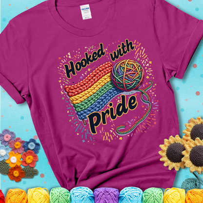 A rainbow-striped crochet swatch paired with a multicolor yarn ball and bold “Hooked With Pride” lettering. Confetti surrounds the vibrant scene showing a magenta tee with a teal sunflower background