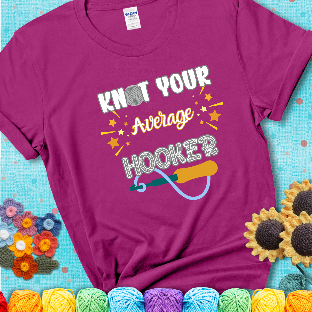 Bold lettering on a magenta t-shirt with a yarn hook graphic. Text reads: “I'm Not Your Average Hooker.” A cheeky crochet tee for confident crafters who own their skills.