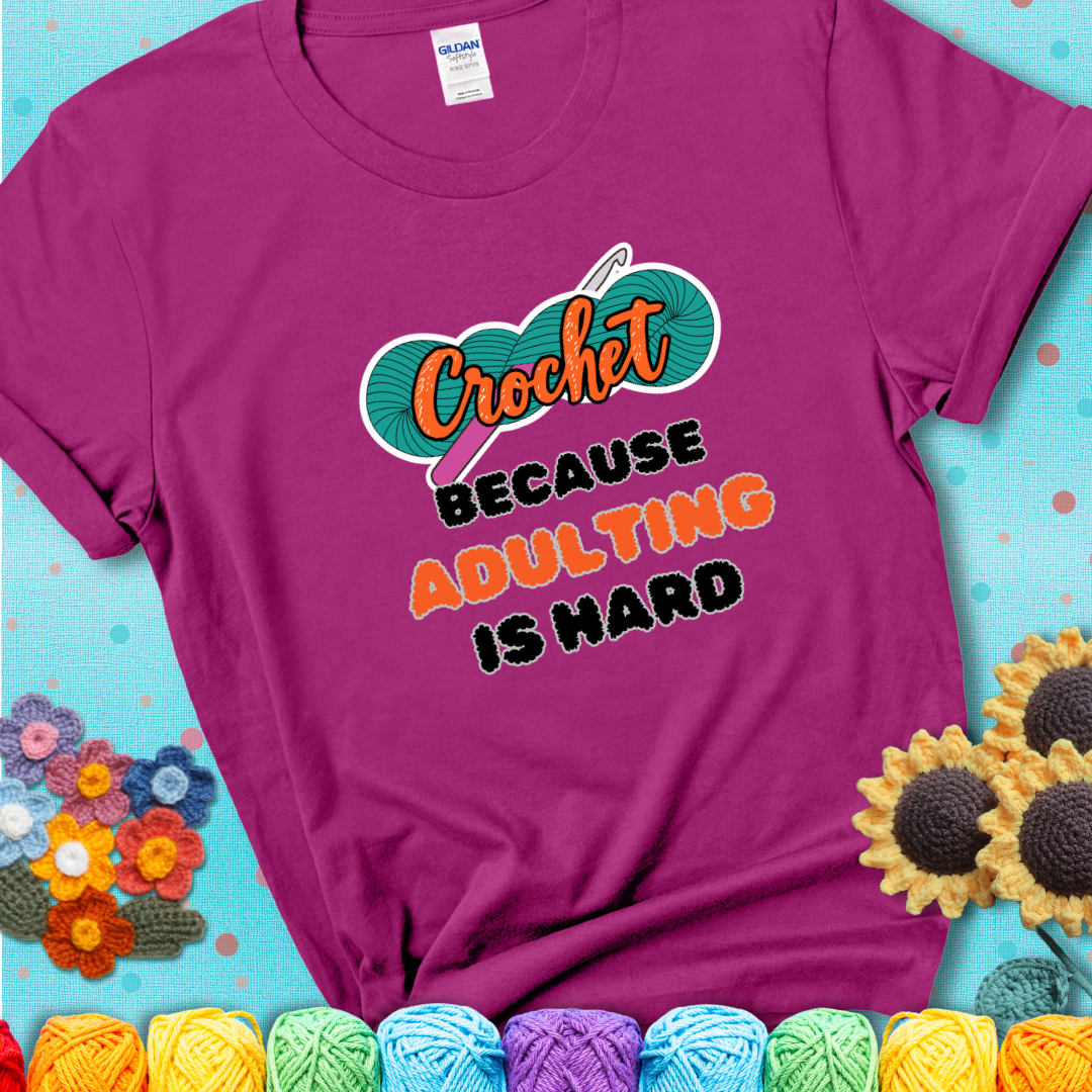 Magenta t-shirt featuring a bold design with the phrase “Crochet Because Adulting Is Hard” featuring a green skein of yarn with a pink crochet hook. The word “Crochet” is in a script font, while “Adulting is hard” is in chunky textured letters.
