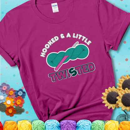 A teal skein of yarn with a hot pink crochet hook through it, captioned “Hooked & a Little Twisted” on a magenta t-shirt.
