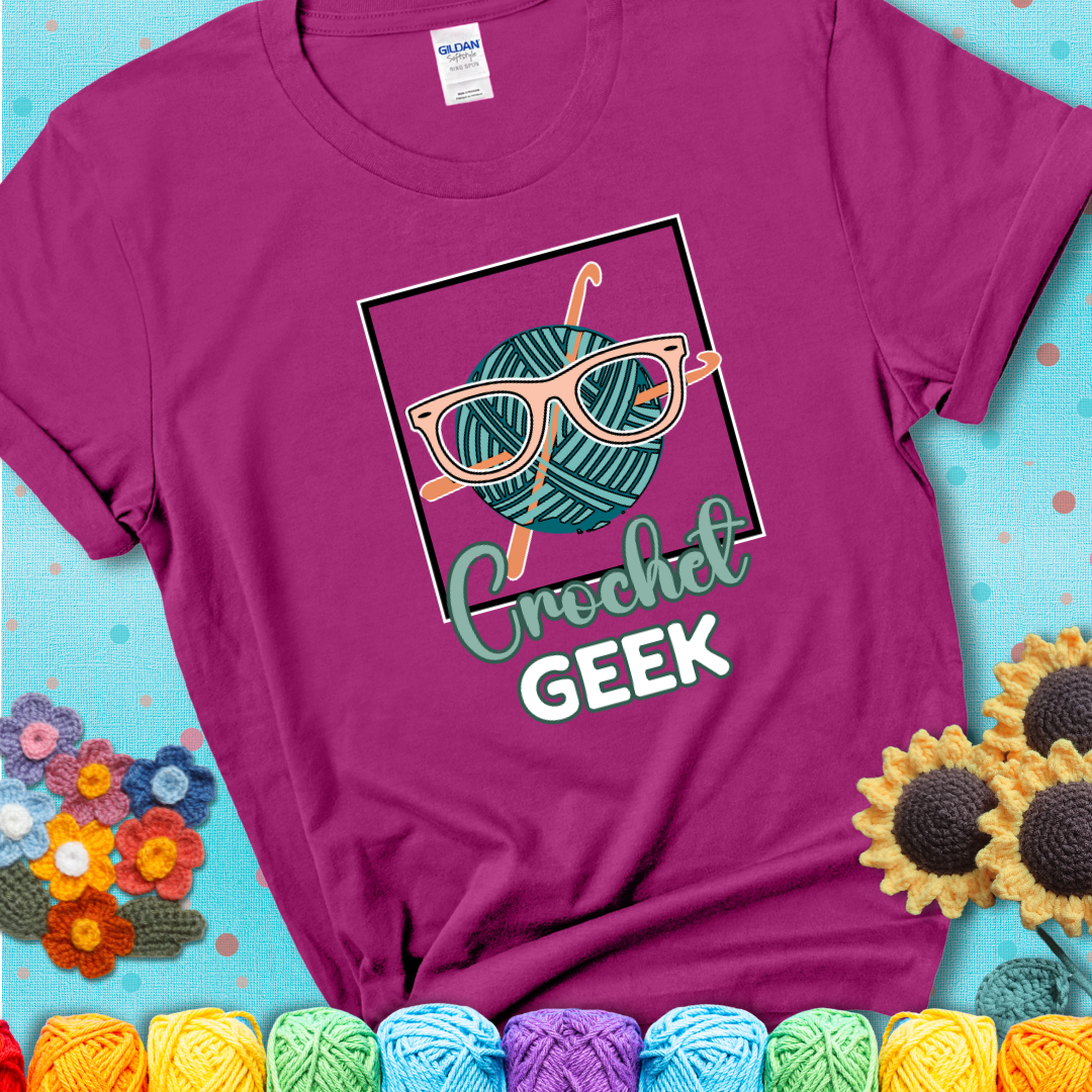A teal yarn ball with crisscrossed hooks wears oversized nerdy glasses with the phrase “Crochet Geek” in bold, fun text below on a magenta t-shirt.