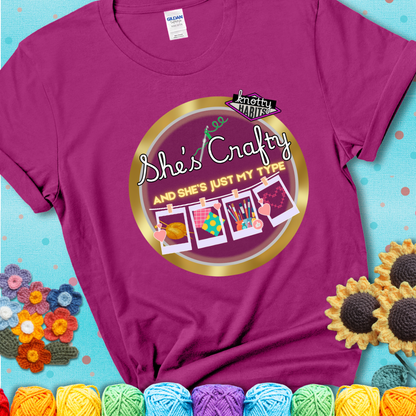 Text on magenta tee reads "She’s Crafty & She’s Just My Type" with yarn, fabric, painting, and cross-stitch icons hanging like Polaroids