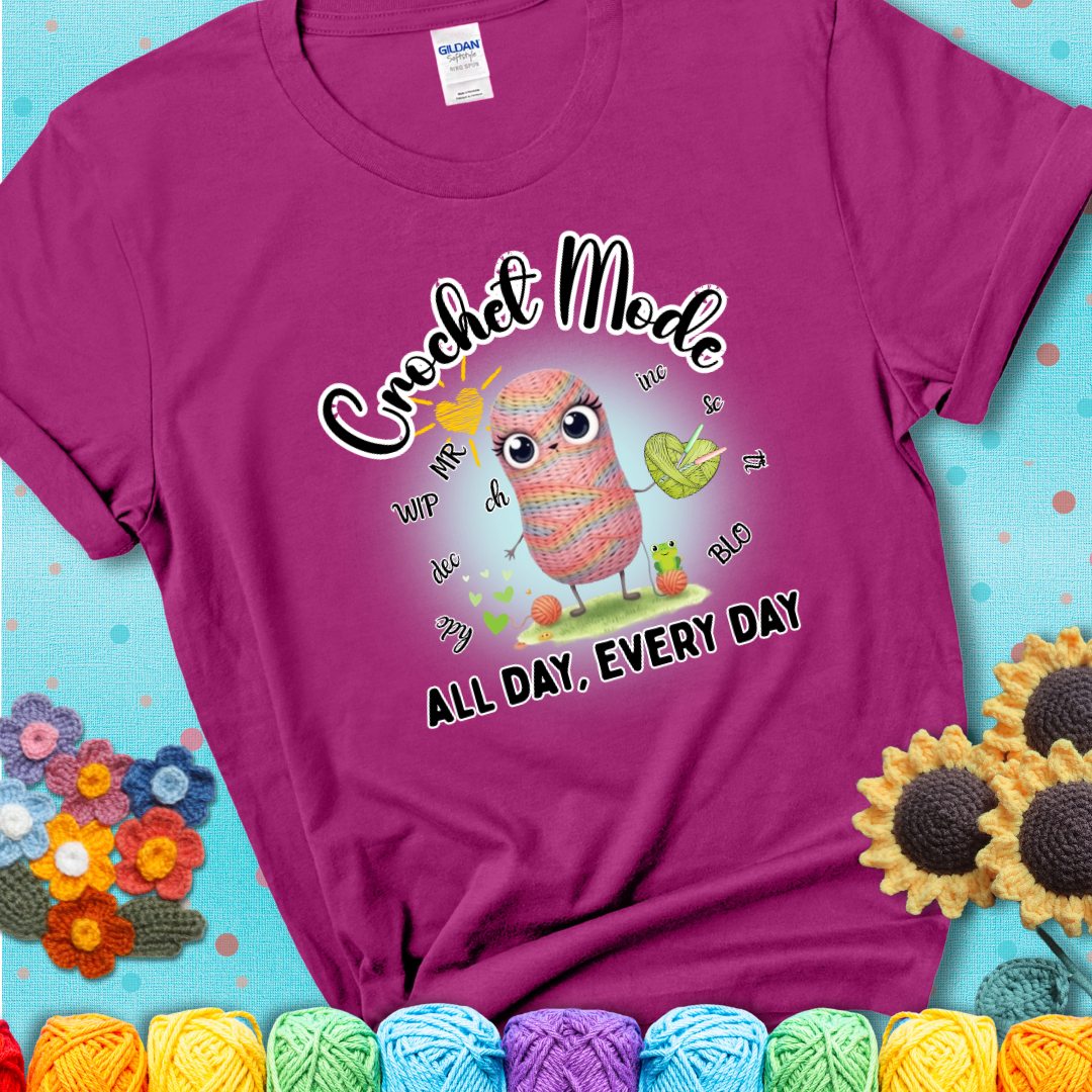 Magenta t-shirt with a kawaii-style rainbow yarn character surrounded by crochet stitch abbreviations and holding a yarn heart, with text “Crochet Mode All Day, Every Day.”
