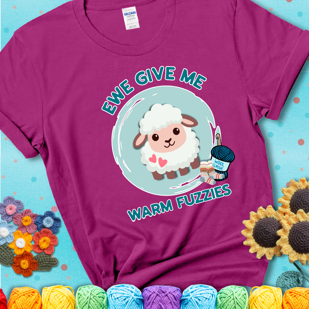 Magenta t-shirt showing a cartoon sheep with pink hearts, wool socks, and yarn labeled “Wool Classic.” Text reads “Ewe Give Me Warm Fuzzies.”