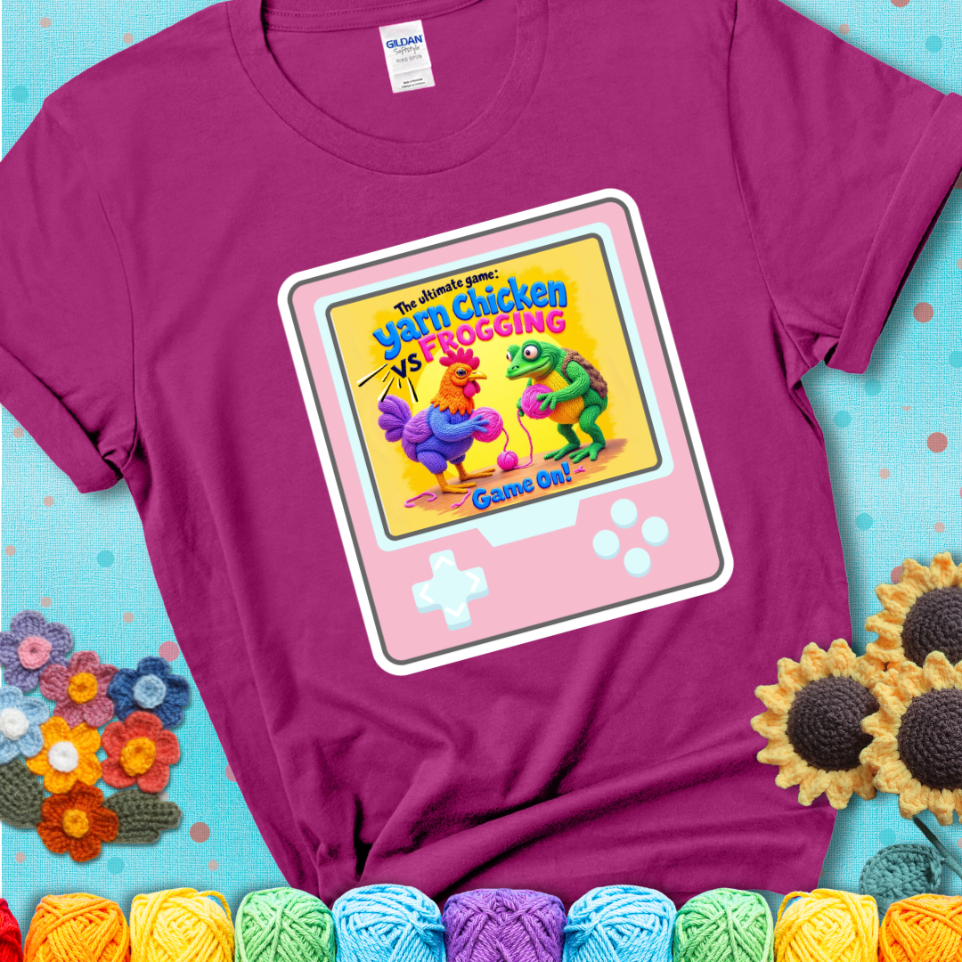 A magenta t-shirt featuring a cartoon yarn chicken and frog face off with text “Yarn Chicken vs Frogging: Game On!” on a retro, handheld video game meets crochet humor battle design.