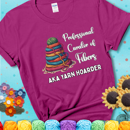 A magenta t-shirt showing a pyramid of rainbow yarn topped with green ball; text reads “Professional Curator of Fibers AKA Yarn Hoarder”, a playful shirt for serious stashers.