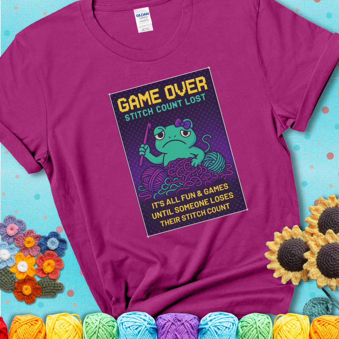 Magenta t-shirt shows a pixel-style design of a grumpy yarn frog holding a hook, surrounded by yarn chaos, with the text “Game Over. Stitch Count Lost.”