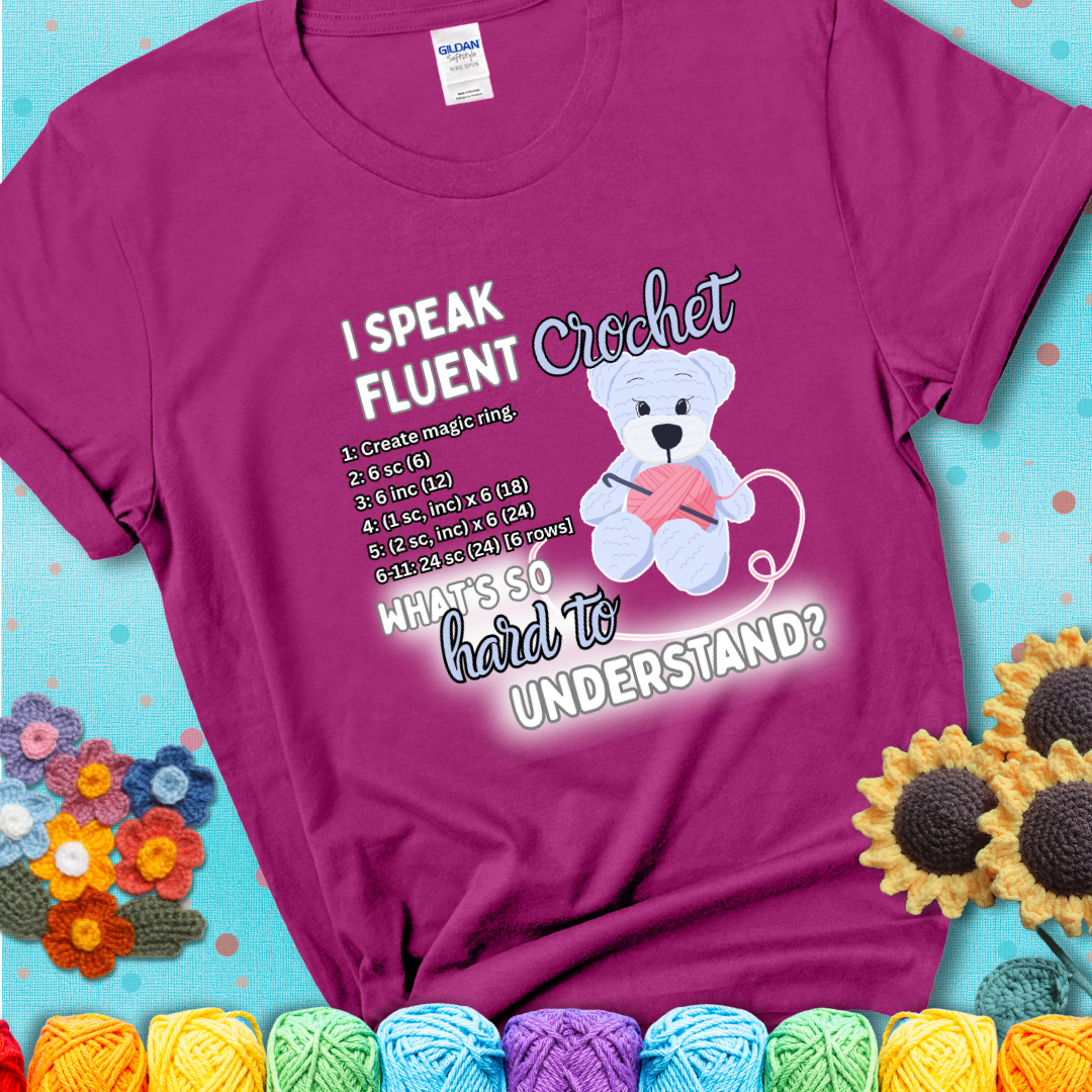 A cute crochet bear holding a yarn ball, surrounded by a short crochet pattern. The phrase reads “I Speak Fluent Crochet – What’s so hard to understand?” on a magenta t-shirt.