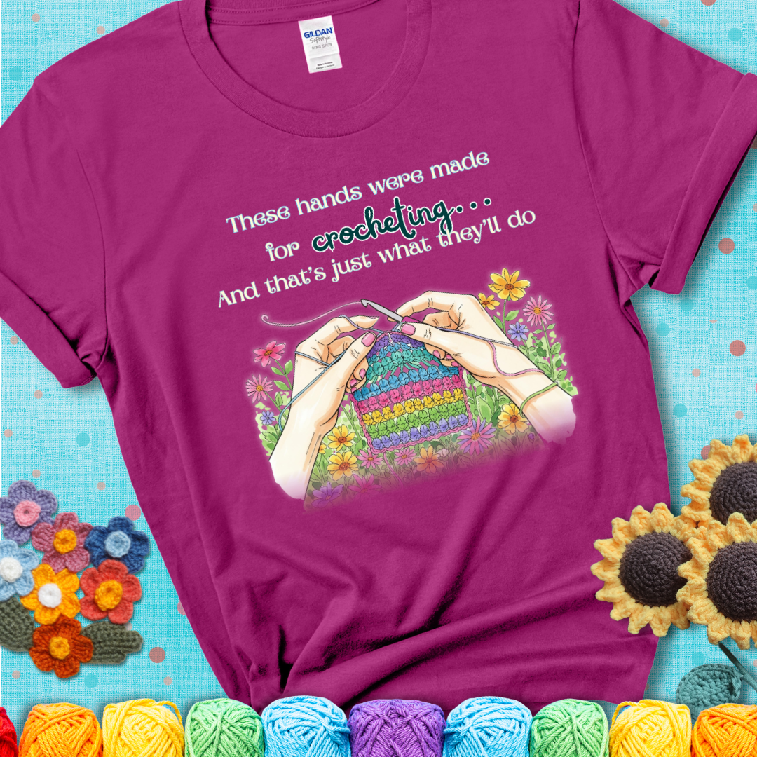 Antique Heliconia t-shirt with an illustration of two hands crocheting a rainbow swatch, surrounded by colorful flowers and the phrase “These hands were made for crocheting… and that’s just what they’ll do.”