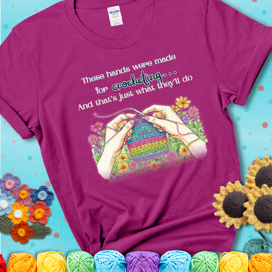Antique Heliconia t-shirt with an illustration of two hands crocheting a rainbow swatch, surrounded by colorful flowers and the phrase “These hands were made for crocheting… and that’s just what they’ll do.”