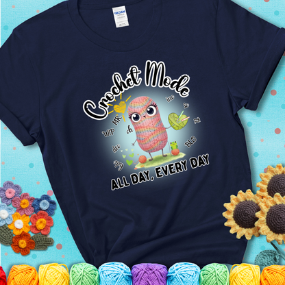 Navy blue t-shirt with a kawaii-style rainbow yarn character surrounded by crochet stitch abbreviations and holding a yarn heart, with text “Crochet Mode All Day, Every Day.”