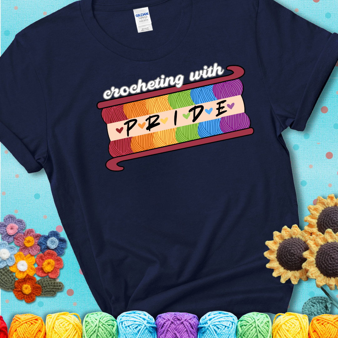 Yarn skeins in rainbow colors stacked in a hook shape. Text reads “Crocheting With Pride” showing a navy blue tee with a teal sunflower background