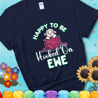 Navy blue t-shirt with smiling cartoon sheep peeks from a pink yarn ball, surrounded by green text “Happy to Be Hooked on Ewe”, a playful and affectionate sheep pun for crocheters.