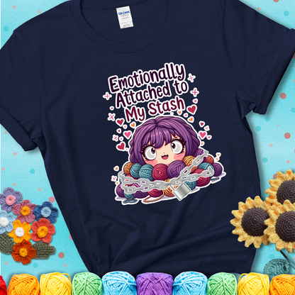 Adorable character with wide eyes hugging a yarn stash wrapped in chains and a lock, with the text “Emotionally Attached to My Stash”  on a navy blue tee.