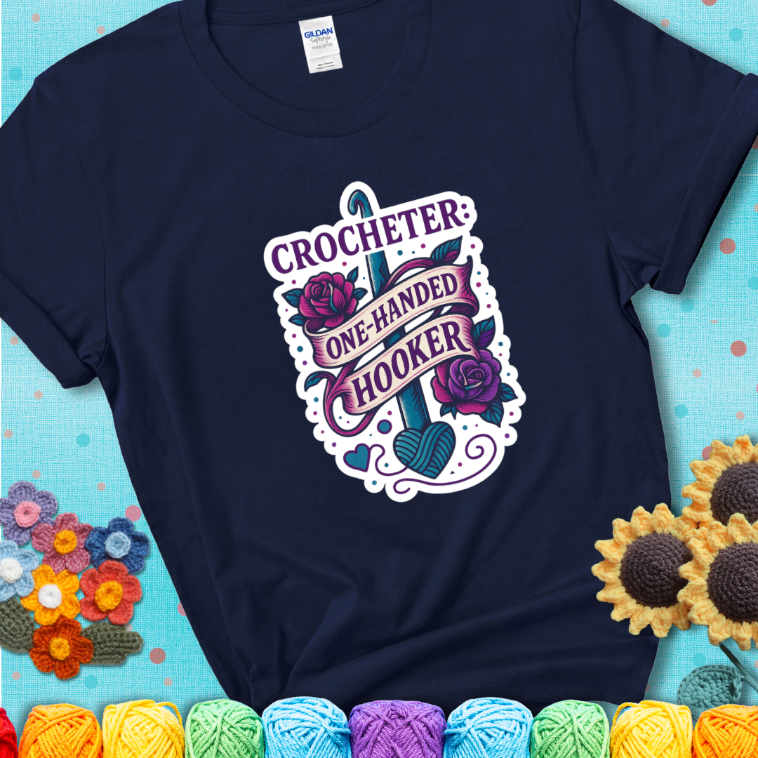 Navy t-shirt with a tattoo-style crochet design featuring a crochet hook, roses, and the phrase “Crocheter: One-Handed Hooker.”