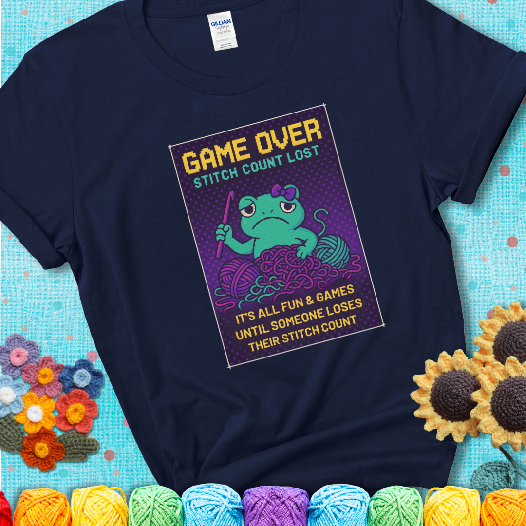 Navy blue t-shirt shows a pixel-style design of a grumpy yarn frog holding a hook, surrounded by yarn chaos, with the text “Game Over. Stitch Count Lost.”