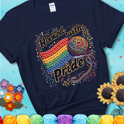 A rainbow-striped crochet swatch paired with a multicolor yarn ball and bold “Hooked With Pride” lettering. Confetti surrounds the vibrant scene showing a navy blue tee with a teal sunflower background