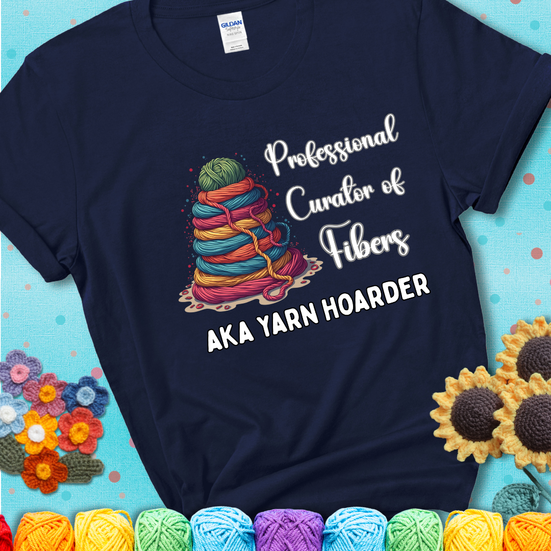 A navy blue t-shirt showing a pyramid of rainbow yarn topped with green ball; text reads “Professional Curator of Fibers AKA Yarn Hoarder”, a playful shirt for serious stashers.