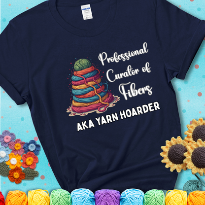A navy blue t-shirt showing a pyramid of rainbow yarn topped with green ball; text reads “Professional Curator of Fibers AKA Yarn Hoarder”, a playful shirt for serious stashers.