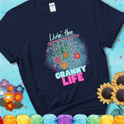 Navy blue t-shirt shows colorful crochet granny squares hanging from a hook with text “Livin’ the Granny Life” in bold pink and blue font; a fun nod to classic crochet.